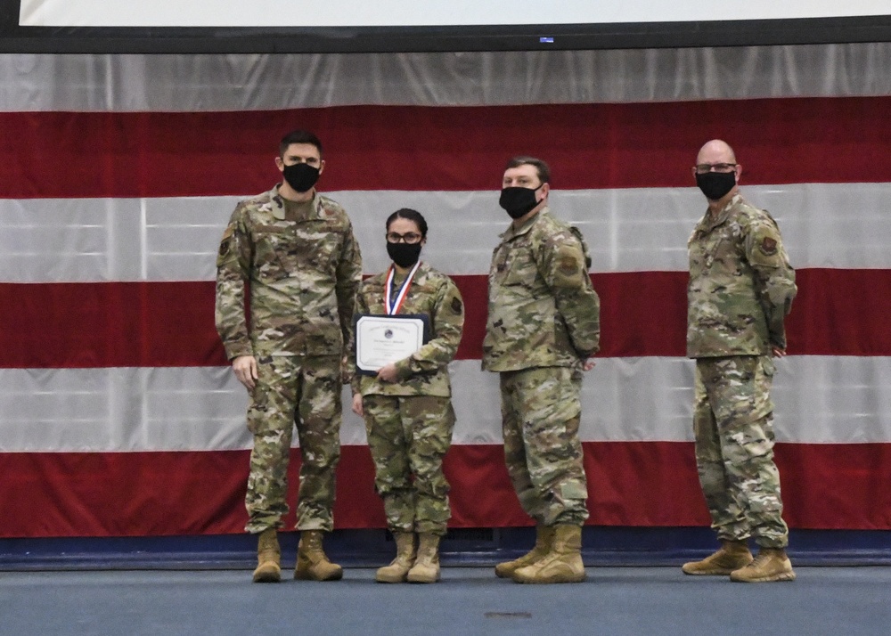 Four military personnel in OCP (Operational Camouflage Pattern) uniforms stand in front of a large American flag backdrop during what appears to be an awards ceremony. One individual, second from left, is holding a certificate and wearing a medal with a red, white, and blue ribbon. All four individuals are wearing black face masks, suggesting this event took place during the COVID-19 pandemic period.
