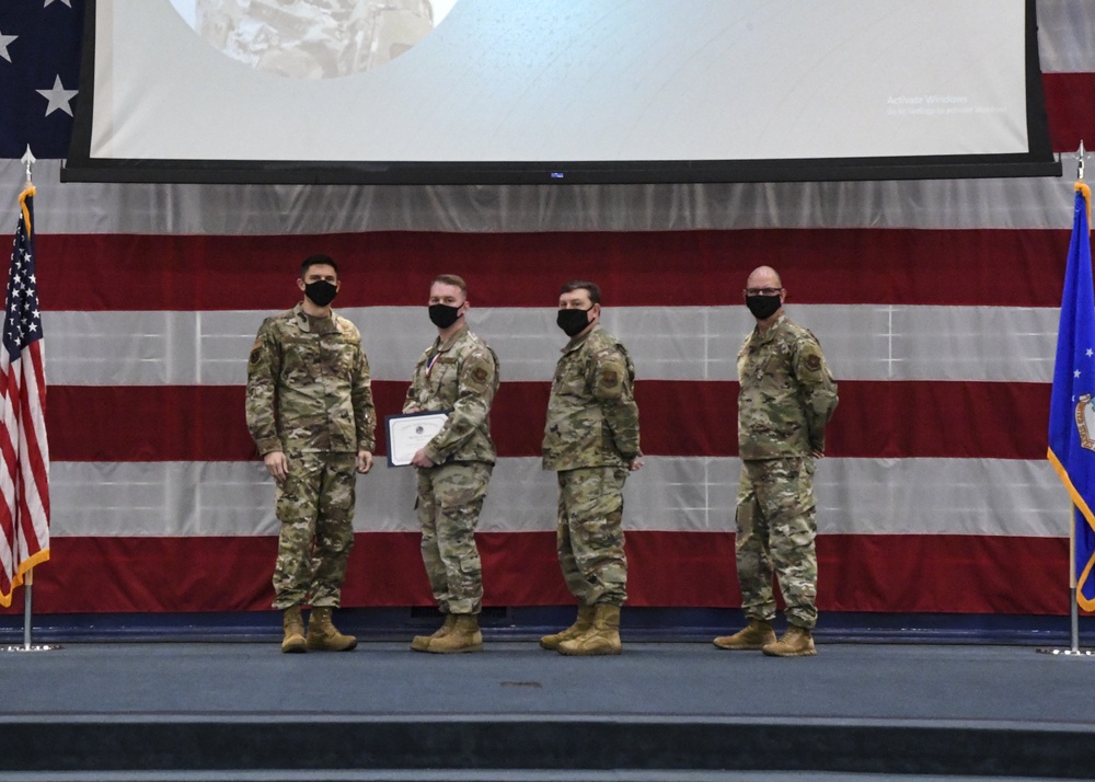 Four military personnel in camouflage uniforms stand on a stage during what appears to be an awards or recognition ceremony. One individual is holding a certificate or award document. The backdrop features a large American flag and a blue Air Force flag, with a projection screen above.