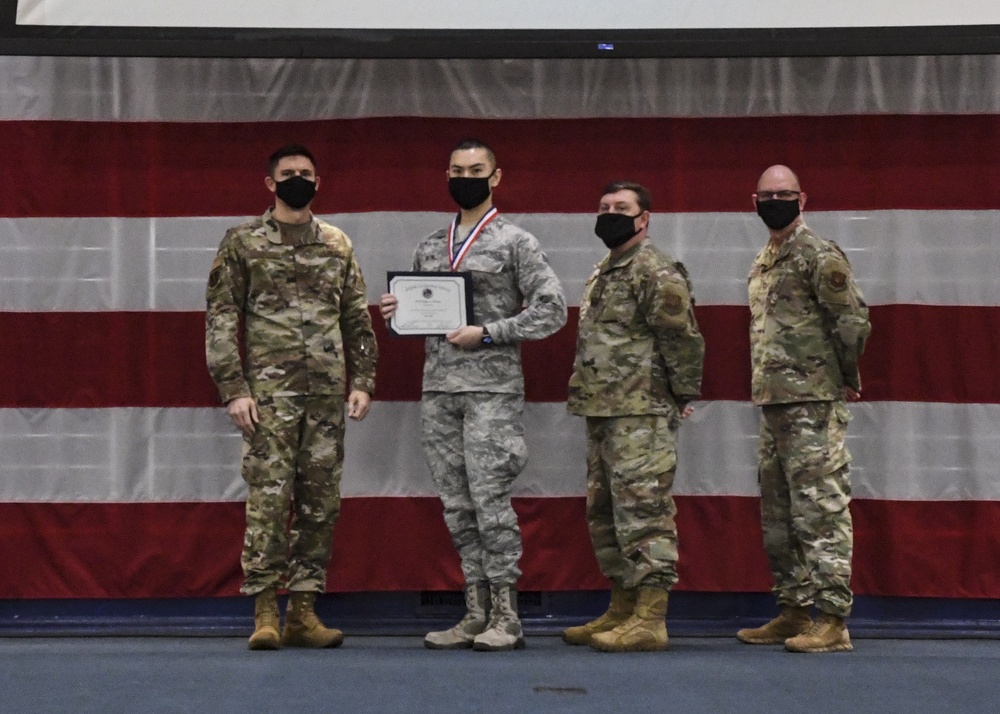 Four military personnel are posed in front of a large American flag backdrop, all wearing black face masks. The second individual from the left is holding a framed certificate and wearing a medal around their neck, suggesting a formal recognition or award ceremony. Three of the four individuals are wearing OCP (Operational Camouflage Pattern) uniforms, while the award recipient appears to be wearing an Airman Battle Uniform (ABU).