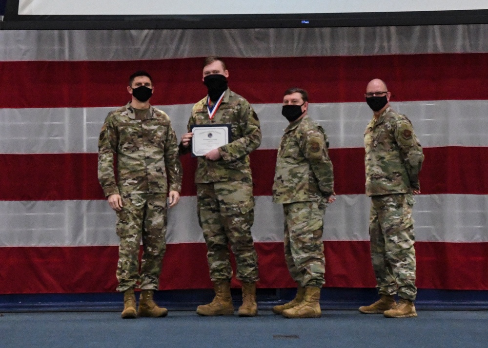 Four military personnel in OCP (Operational Camouflage Pattern) uniforms pose for a formal photo in front of a large American flag backdrop. The second individual from the left is holding a certificate and wearing a medal around their neck, indicating a military award or recognition ceremony. All four individuals are wearing black face masks, suggesting this event took place during the COVID-19 pandemic period.