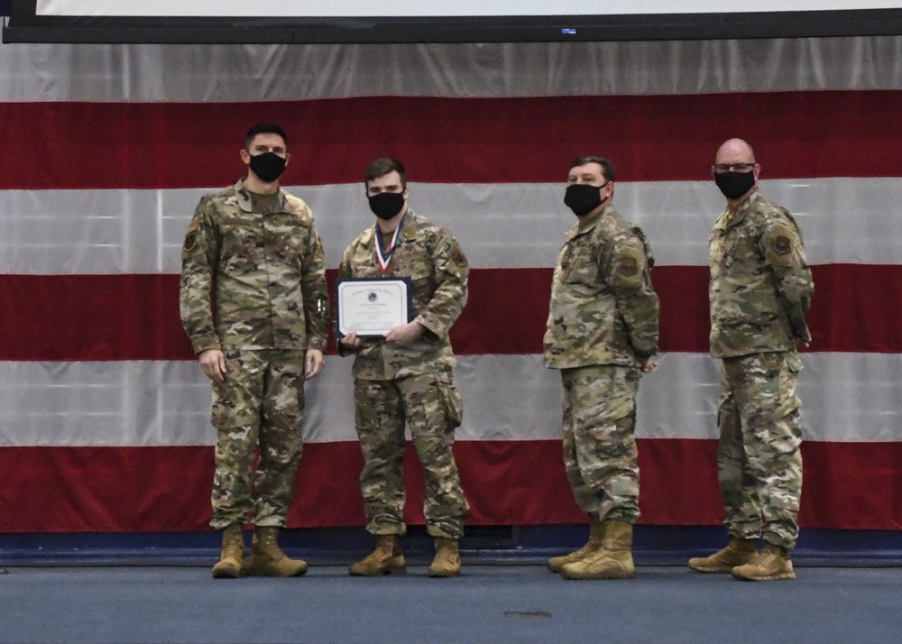 Four military personnel in OCP (Operational Camouflage Pattern) uniforms pose in front of a large American flag backdrop. One individual, wearing a medal around their neck, holds a framed certificate or award document. All four individuals are wearing black face masks, suggesting this ceremony took place during the COVID-19 pandemic period.