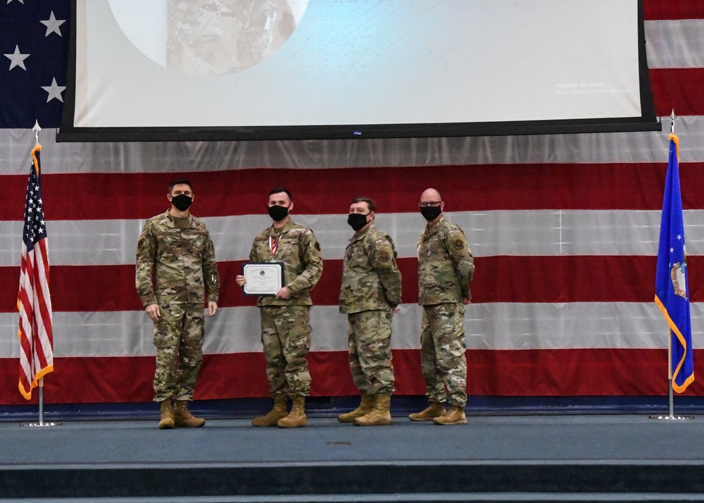Four military personnel in OCP (Operational Camouflage Pattern) uniforms stand on a stage during a formal awards ceremony. One individual is holding a certificate and appears to be wearing a medal, suggesting a recognition or commendation event. All four are wearing black face masks, consistent with COVID-19 era protocols.