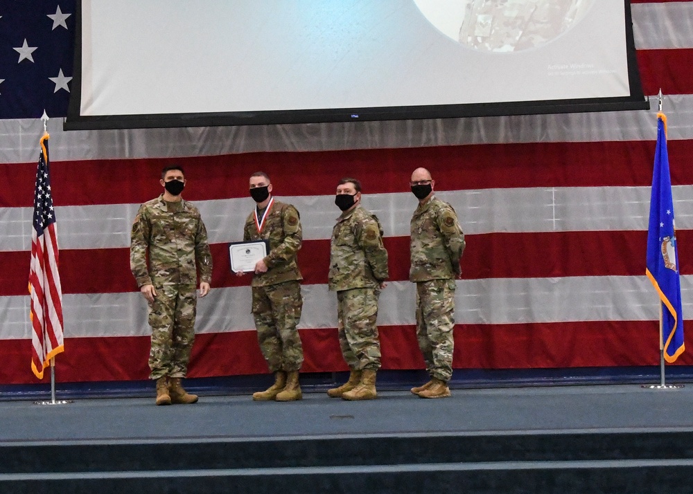 Four military personnel in OCP (Operational Camouflage Pattern) uniforms stand on a stage during what appears to be an awards or recognition ceremony. One individual is wearing a medal around their neck and holding a certificate, which is being presented by another service member. The backdrop features a large American flag and a projection screen, with a US Air Force flag visible on the right side of the stage.
