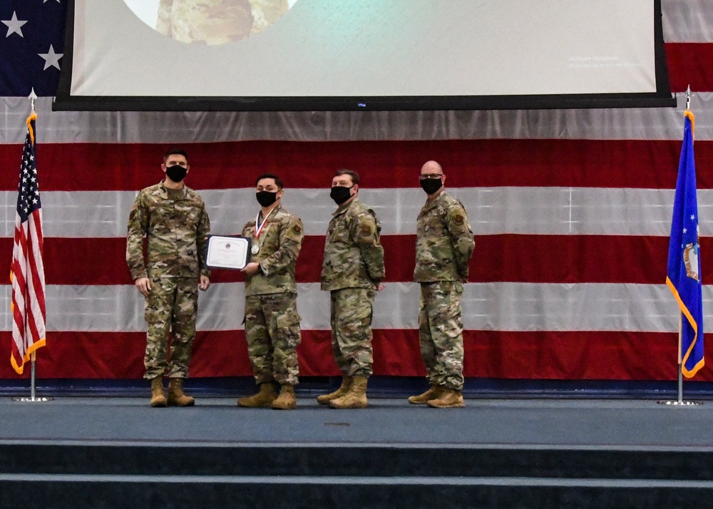Four military personnel in OCP (Operational Camouflage Pattern) uniforms stand on a stage during a formal awards ceremony. One service member is holding a certificate and wearing a medal, indicating they are the recipient of a commendation or award. All individuals are wearing black face masks, suggesting the event took place during the COVID-19 pandemic period.