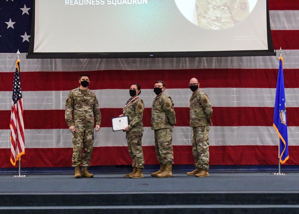 Four military personnel in OCP (Operational Camouflage Pattern) uniforms stand on a stage during what appears to be an awards or recognition ceremony. One individual is holding a certificate and wearing a medal or ribbon around their neck. The setting includes a large American flag backdrop, a projection screen displaying 'Readiness Squadron,' and flanking flags including a US flag and what appears to be an Air Force flag.