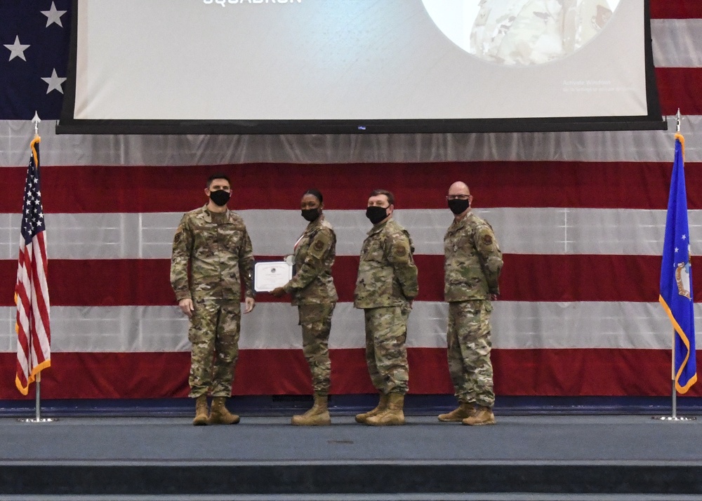 Four U.S. Air Force personnel in OCP (Operational Camouflage Pattern) uniforms stand on a stage during a formal military ceremony. One individual is receiving or holding a certificate/award, suggesting this is a recognition or awards ceremony. All four individuals are wearing black face masks, indicating this event took place during the COVID-19 pandemic period.