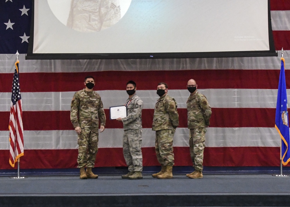 Four military personnel in camouflage uniforms and black face masks are posed on a stage during a formal ceremony. Two individuals in the center are jointly holding a certificate or award document. The backdrop features a large American flag drape, a projection screen, and flanking flags including the US flag and what appears to be an Air Force flag.