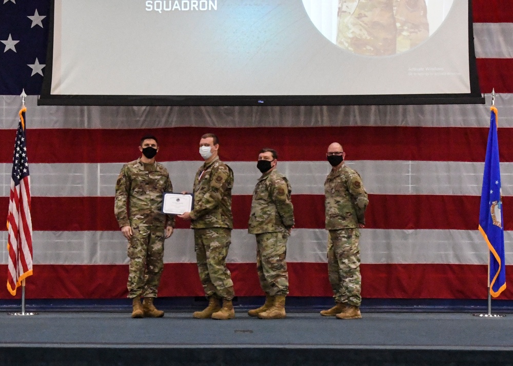 Four military personnel in OCP (Operational Camouflage Pattern) uniforms stand on a stage during a formal ceremony, with two individuals holding a certificate or award document. All four are wearing black face masks, suggesting this event took place during the COVID-19 pandemic period. The backdrop features a large American flag drape and a projection screen displaying the word 'SQUADRON'.