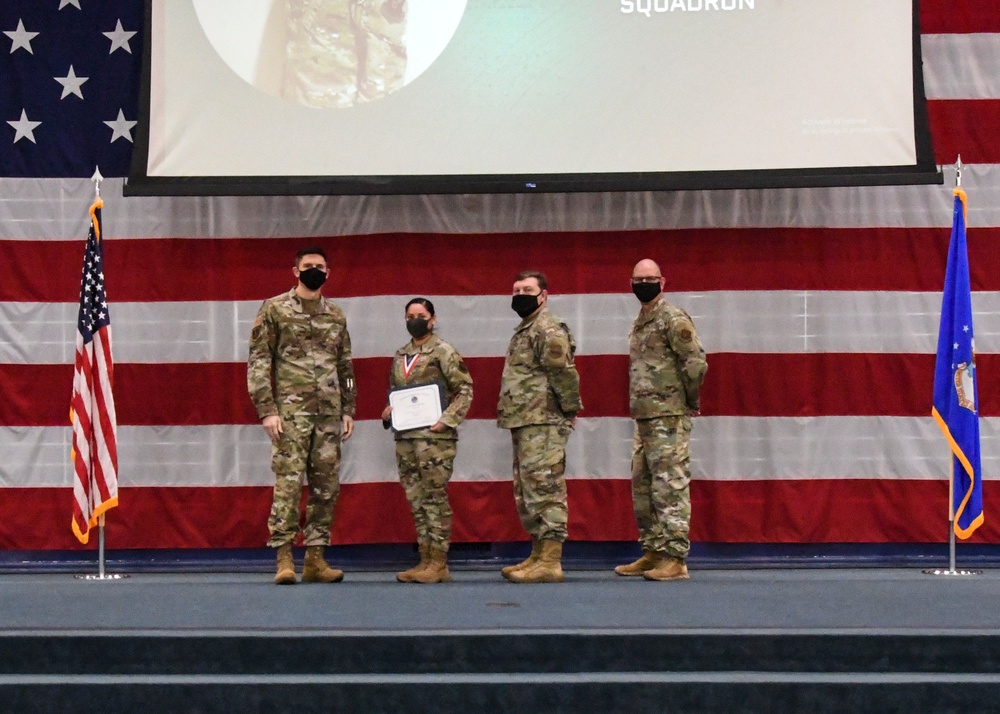 Four military personnel in OCP (Operational Camouflage Pattern) uniforms stand on a stage during what appears to be an awards ceremony. One individual, likely female, is holding a certificate and wearing a medal around their neck. The backdrop features a large American flag and a projection screen displaying the word 'SQUADRON'.