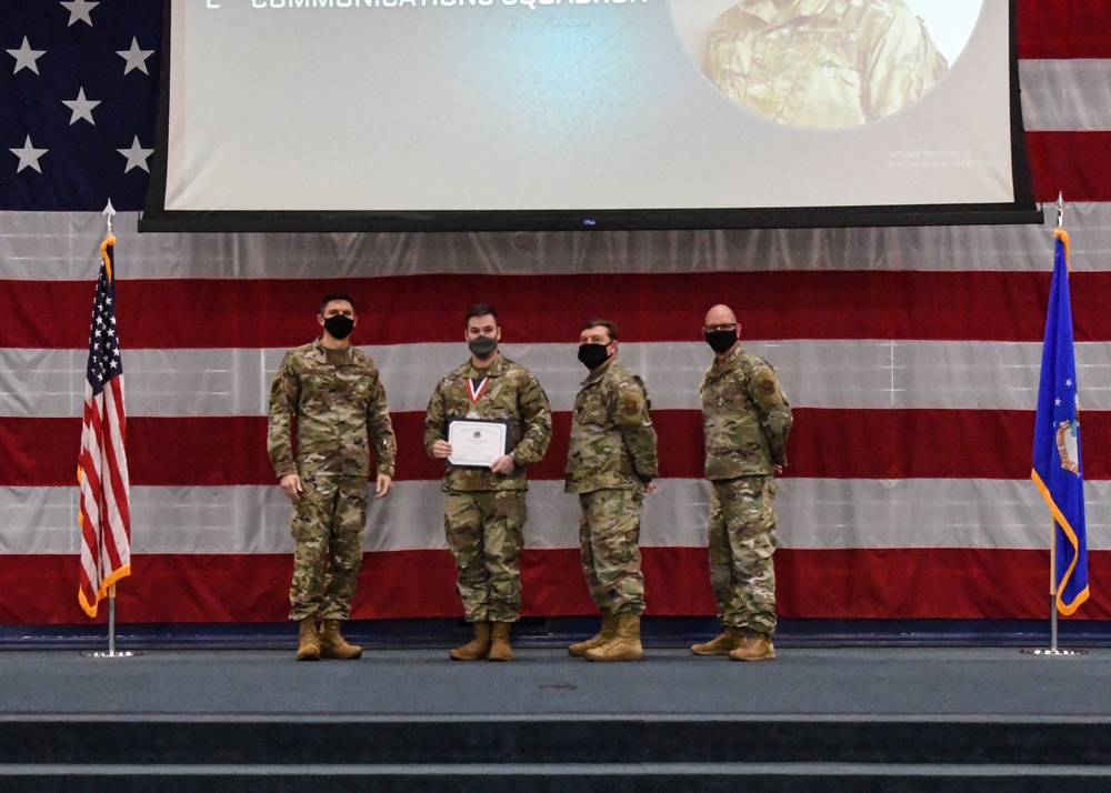 Four military personnel in camouflage uniforms and face masks are posed on a stage during an awards ceremony. One individual in the center is holding a certificate and wearing a medal around their neck. The backdrop features a large American flag and a projection screen displaying text referencing a 'Communications Squadron.'