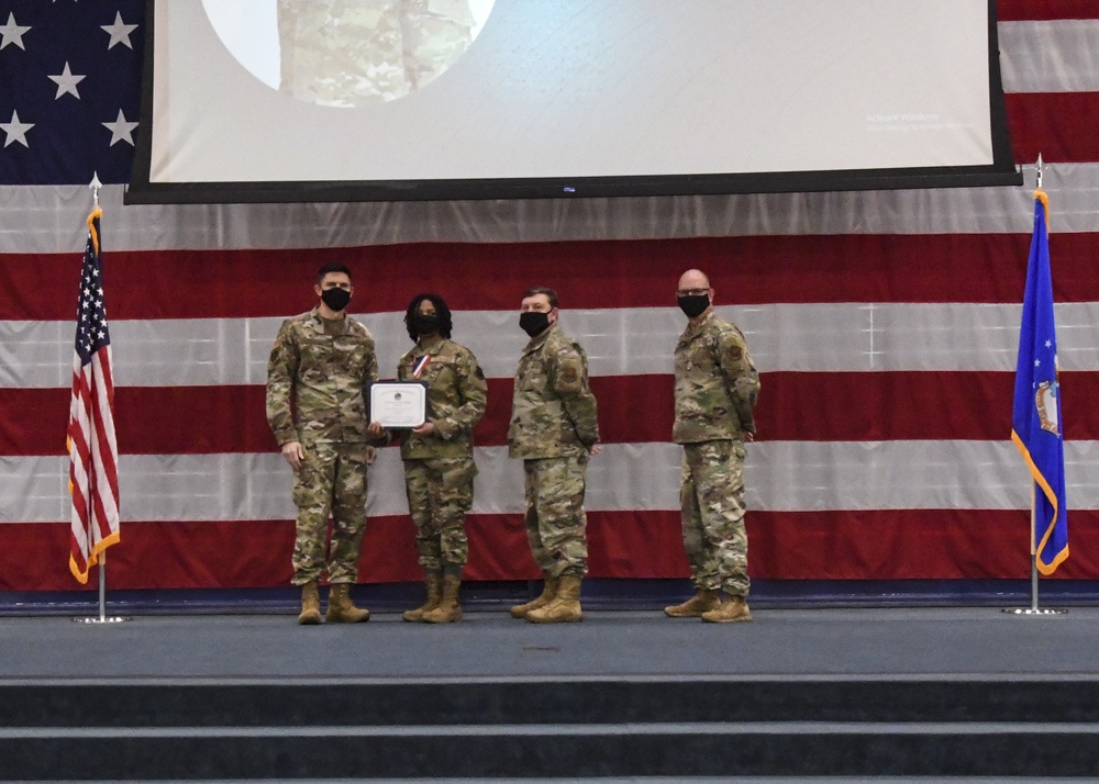 Four military personnel in camouflage uniforms stand on a stage during what appears to be an awards or recognition ceremony. One individual is holding a certificate and wearing a medal, suggesting they are the recipient of an award. All four individuals are wearing black face masks, indicating this ceremony took place during the COVID-19 pandemic period.