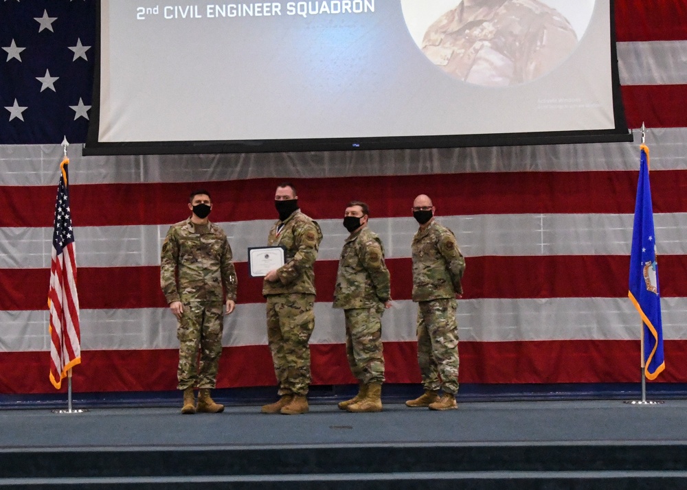 Four military personnel in OCP (Operational Camouflage Pattern) uniforms stand on a stage during a formal ceremony for the 2nd Civil Engineer Squadron. Two individuals in the center are exchanging or holding a certificate/award together, while all four wear black face masks. The backdrop features a large American flag drape, a projection screen displaying '2nd Civil Engineer Squadron,' and flanking flags including a US flag and what appears to be an Air Force flag.