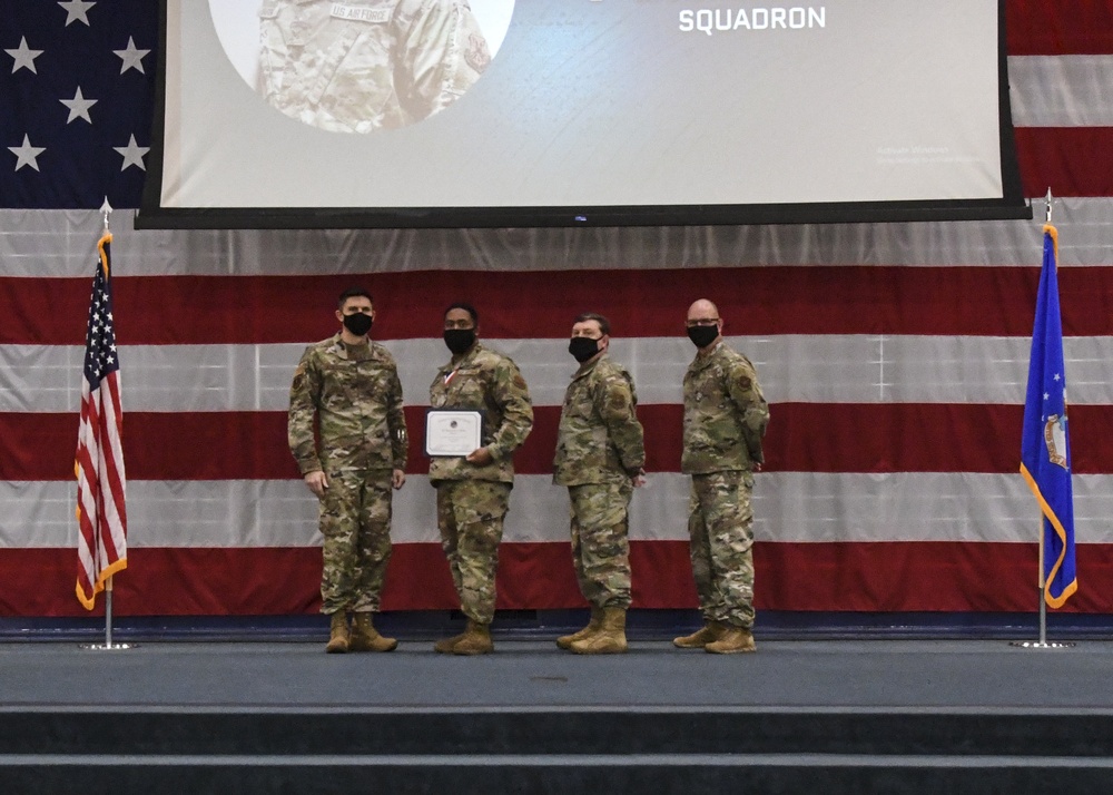 Four U.S. Air Force personnel in OCP (Operational Camouflage Pattern) uniforms stand on a stage during a military awards ceremony. One service member is holding a certificate/award while wearing a medal, suggesting a formal recognition event. All four individuals are wearing black face masks, indicating this ceremony took place during the COVID-19 pandemic period.