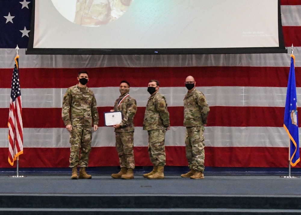 Four military personnel in OCP (Operational Camouflage Pattern) uniforms stand on a stage during a formal awards ceremony. One individual is holding a certificate and appears to be wearing a medal, suggesting a commendation or achievement recognition event. All four individuals are wearing black face masks, indicating this ceremony took place during the COVID-19 pandemic period.