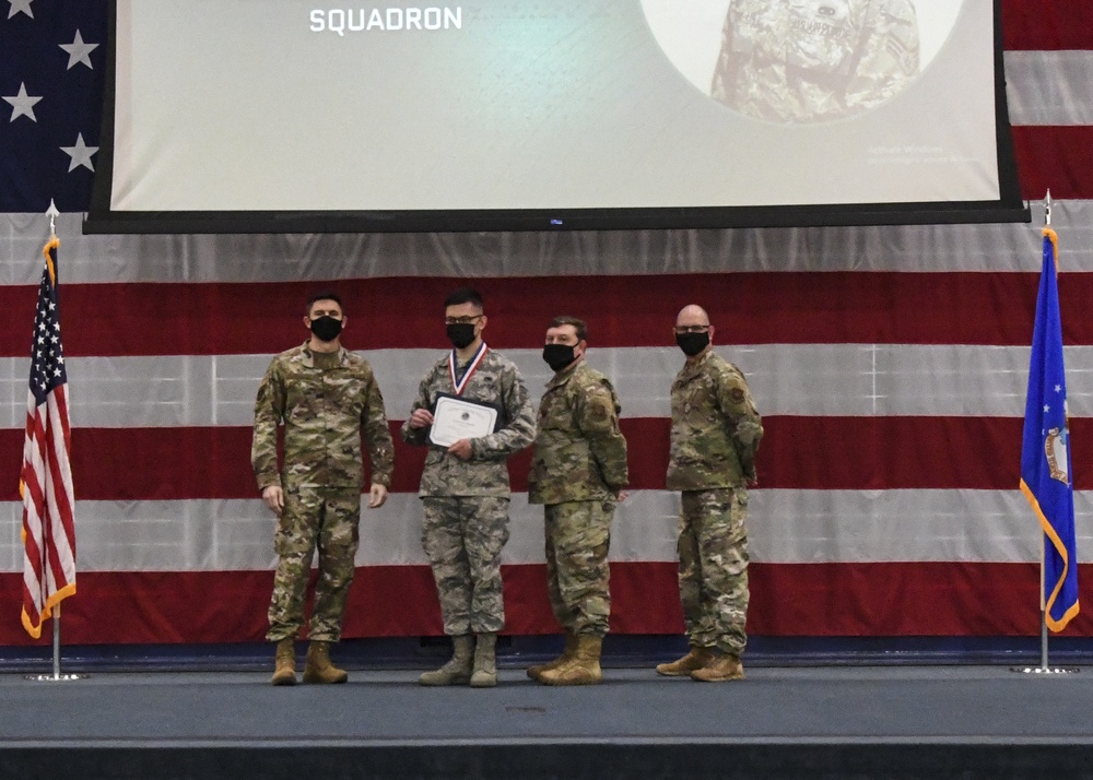 Four military personnel in OCP (Operational Camouflage Pattern) uniforms are posed on a stage during an awards ceremony. One individual is holding a certificate and wearing a medal around their neck, indicating a formal recognition event. The backdrop features a large American flag and a projection screen displaying the word 'SQUADRON' along with a military emblem.