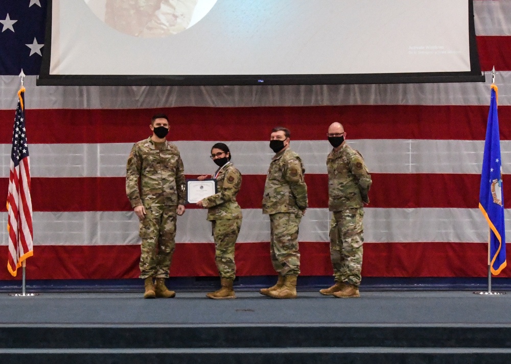 Four military personnel in OCP (Operational Camouflage Pattern) uniforms are posed on a stage during a formal ceremony. One service member is receiving a certificate or award, being presented by another member while two others stand at attention. The backdrop features a large American flag drape and a projection screen, with the US flag and what appears to be an Air Force flag flanking the stage.