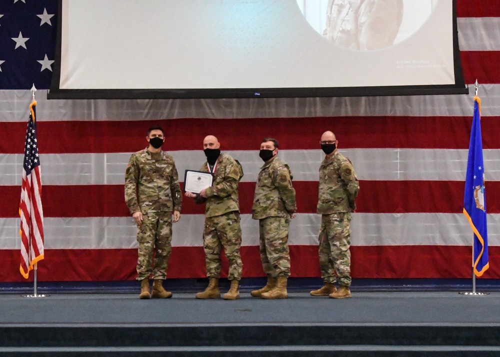 Four military personnel in OCP (Operational Camouflage Pattern) uniforms stand on a stage during a formal awards or recognition ceremony. Two individuals in the center are jointly holding a certificate or award document. All four are wearing black face masks, suggesting this event took place during the COVID-19 pandemic period.