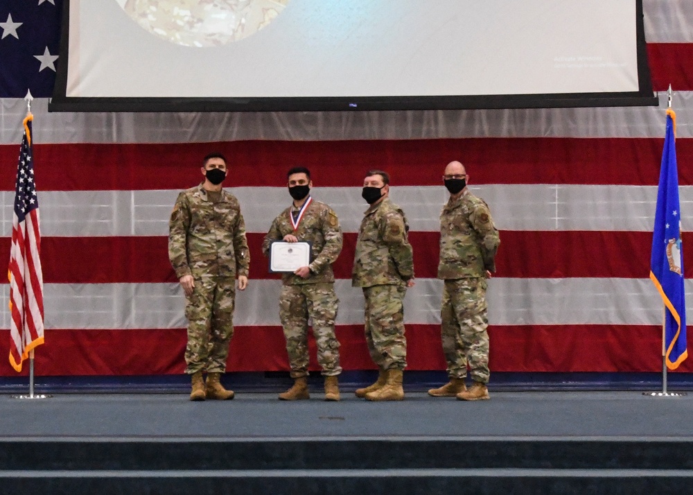 Four military personnel in OCP (Operational Camouflage Pattern) uniforms stand on a stage during a formal awards ceremony. One individual in the center is holding a certificate and wearing a medal around their neck, indicating they are the recipient of a commendation or award. All four individuals are wearing black face masks, suggesting the event took place during the COVID-19 pandemic period.