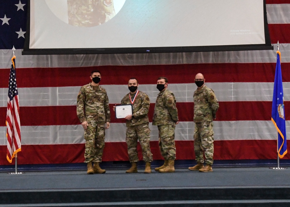 Four military personnel in OCP (Operational Camouflage Pattern) uniforms stand on a stage during a formal awards ceremony. One individual, wearing a medal around their neck, is holding a certificate or award document. The backdrop features a large American flag drape and a projection screen, with an Air Force flag visible on the right side.