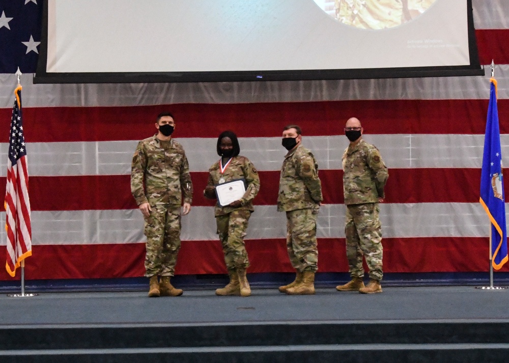 Four military personnel in OCP (Operational Camouflage Pattern) uniforms are standing on a stage during what appears to be an awards ceremony. One individual is holding a certificate and wearing a medal around their neck, indicating they are the recipient of a commendation. The backdrop features a large American flag and a projection screen, with a US Air Force flag visible on the right side of the stage.