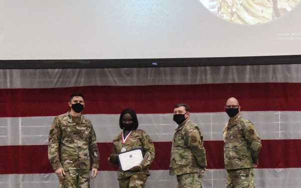 Four military personnel in OCP (Operational Camouflage Pattern) uniforms are standing on a stage during what appears to be an awards ceremony. One individual is holding a certificate and wearing a medal around their neck, indicating they are the recipient of a commendation. The backdrop features a large American flag and a projection screen, with a US Air Force flag visible on the right side of the stage.