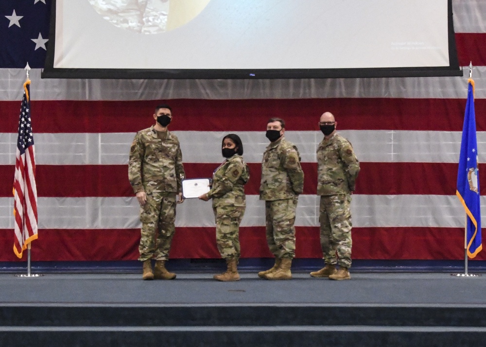 Four military personnel in OCP (Operational Camouflage Pattern) uniforms are posed on a stage during a formal ceremony. A female service member is receiving or presenting a certificate/award document to another service member, flanked by two others. The backdrop features a large American flag and a projected screen, with a US Air Force flag visible on the right.