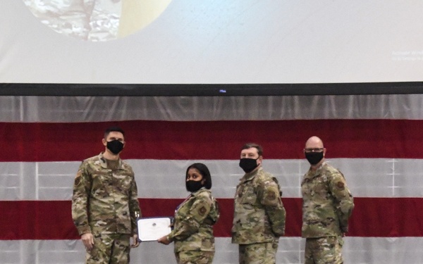 Four military personnel in OCP (Operational Camouflage Pattern) uniforms are posed on a stage during a formal ceremony. A female service member is receiving or presenting a certificate/award document to another service member, flanked by two others. The backdrop features a large American flag and a projected screen, with a US Air Force flag visible on the right.
