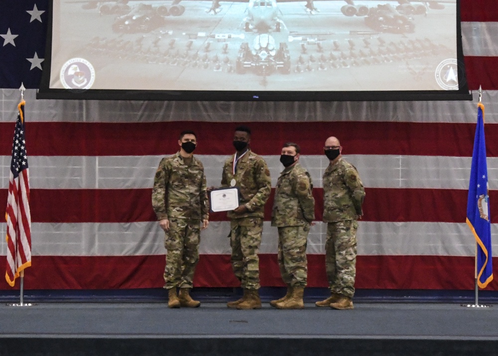 Four military personnel in OCP (Operational Camouflage Pattern) uniforms stand on a stage during a formal awards ceremony. One service member is receiving a certificate and medal while the others present it, all wearing black face masks. The backdrop features a large American flag drape, a projection screen displaying a military aircraft photo, and a blue Air Force flag.