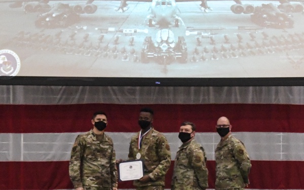 Four military personnel in OCP (Operational Camouflage Pattern) uniforms stand on a stage during a formal awards ceremony. One service member is receiving a certificate and medal while the others present it, all wearing black face masks. The backdrop features a large American flag drape, a projection screen displaying a military aircraft photo, and a blue Air Force flag.