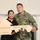 A couple poses together holding a house-shaped wooden sign celebrating a home purchase. The man is wearing a U.S. Army uniform with the name tape 'Thompson' visible, and the woman is dressed in black. The sign reads 'CONGRATS! Sold by Kathy' with a social media handle '@adamshomes' visible.