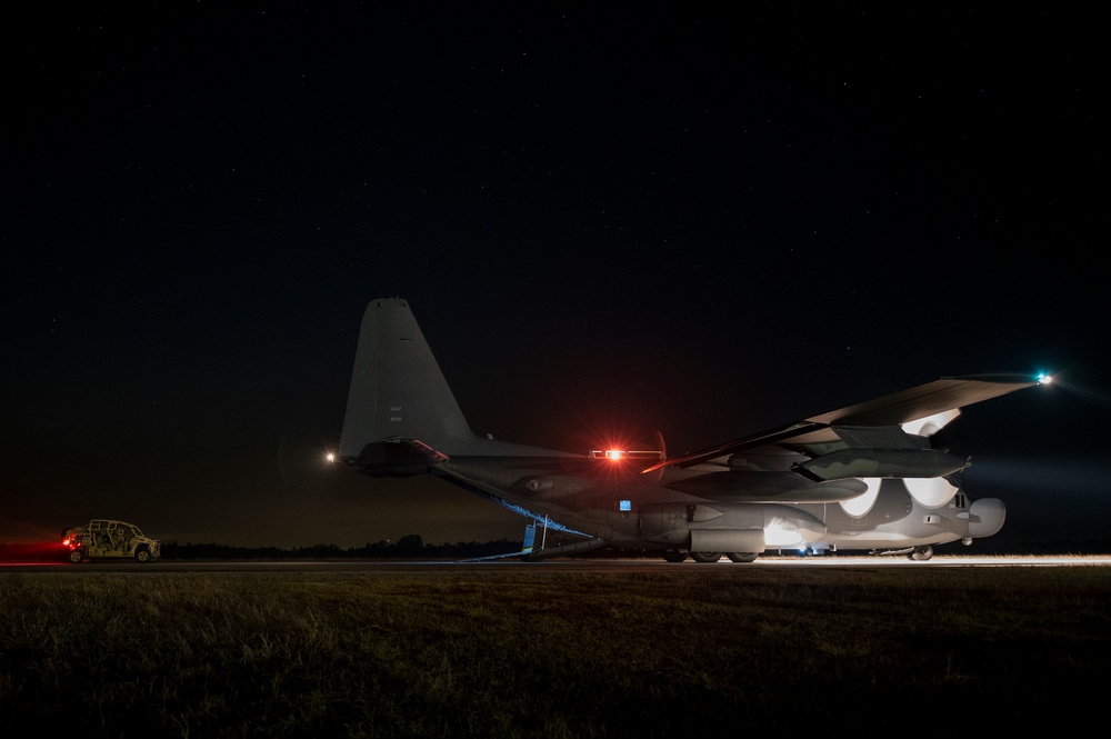 A large military transport aircraft, consistent with a C-130 Hercules variant, is photographed on a runway or taxiway at night under a clear, star-filled sky. The aircraft appears to have its rear cargo ramp lowered, with a military ground vehicle (likely a HMMWV/Humvee) visible to the left. Navigation lights and other illumination sources are active, suggesting the aircraft is powered and potentially conducting loading or unloading operations.