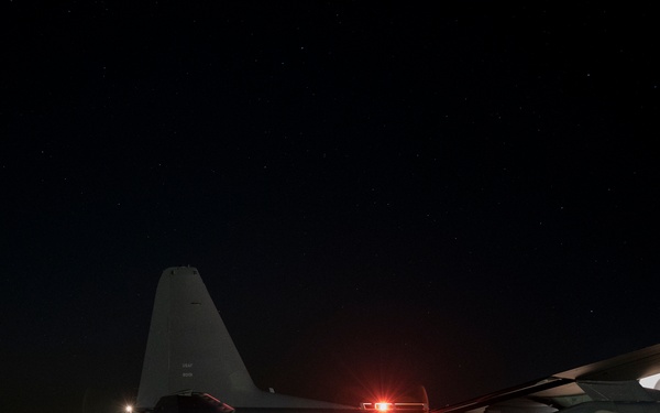 A large military transport aircraft, consistent with a C-130 Hercules variant, is photographed on a runway or taxiway at night under a clear, star-filled sky. The aircraft appears to have its rear cargo ramp lowered, with a military ground vehicle (likely a HMMWV/Humvee) visible to the left. Navigation lights and other illumination sources are active, suggesting the aircraft is powered and potentially conducting loading or unloading operations.