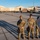 Nellis Weapons Load Crew of the Year Competition Military personnel in OCP (Operational Camouflage Pattern) uniforms are gathered on a flight line apron at what appears to be Nellis Air Force Base, indicated by the 'WA' tail codes visible on the aircraft. Multiple fighter jets, including an F-15 and F-16s with distinctive aggressor-style camouflage paint schemes, are displayed on the tarmac. The scene appears to be a formal static display or ceremony event with large groups of airmen assembled in formation.