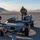 Nellis Weapons Load Crew of the Year Competition Three military personnel in camouflage uniforms and face masks are working on a munitions cart on an airfield tarmac. The cart, labeled 'GZ411' and marked with an 'EXPLOSIVES 1.1' hazard placard, carries what appear to be air-delivered munitions or missiles. Mountains and a military aircraft taking off are visible in the background, suggesting a desert air base environment.