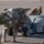 Nellis Weapons Load Crew of the Year Competition Military personnel are operating an MJ-1C munitions lift truck (designated J750) on an airfield tarmac. Two individuals in camouflage uniforms are handling what appears to be an air-to-air missile or similar munition on the loader. The scene appears to be a weapons loading or maintenance operation conducted during early morning or late afternoon light.