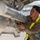 Nellis Weapons Load Crew of the Year Competition A U.S. Air Force airman wearing an OCP (Operational Camouflage Pattern) uniform with yellow reflective safety straps is performing maintenance or inspection work on an aircraft component, likely underneath a military jet. The airman, whose name tape reads 'TWINE,' is closely examining and handling what appears to be a missile or ordnance mounted under the aircraft's wing or fuselage. The image is taken on a flight line or airfield ramp in what appears to be a cold or overcast environment.