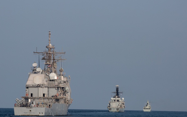 Three naval warships are underway in formation on open blue ocean waters under a hazy gray sky. The closest vessel, prominently visible in the foreground, bears the name 'PORT ROYAL' on its hull and is identifiable as a US Navy Ticonderoga-class guided-missile cruiser. Two additional warships of similar military configuration are visible in the background at increasing distances.