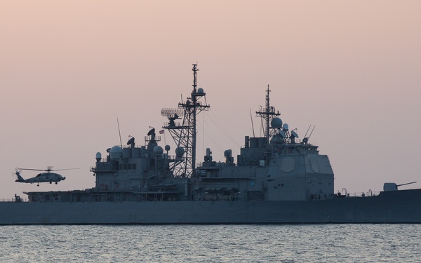 A large U.S. Navy warship, identifiable as a Ticonderoga-class guided-missile cruiser, is photographed at anchor or slow transit on calm waters during twilight or dusk conditions. A military helicopter, consistent with an SH-60 Seahawk, is visible hovering or departing near the ship's port side. The hull number on the bow appears to be '7' followed by additional digits, though full identification is limited by lighting conditions.