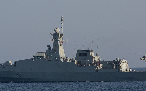 A gray naval warship is underway on open blue water, with a helicopter flying nearby off its stern. The vessel appears to be a modern frigate or corvette with Arabic numerals visible on its hull. The scene suggests a naval exercise or patrol operation.