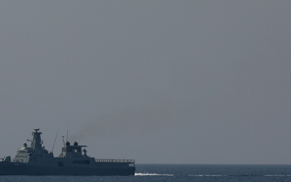 A naval warship is underway at sea with a military jet aircraft flying overhead in the upper right portion of the frame. The vessel appears to be a modern frigate or destroyer with visible superstructure, radar systems, and weapons mounts. The scene suggests a joint naval and air force exercise or patrol operation.