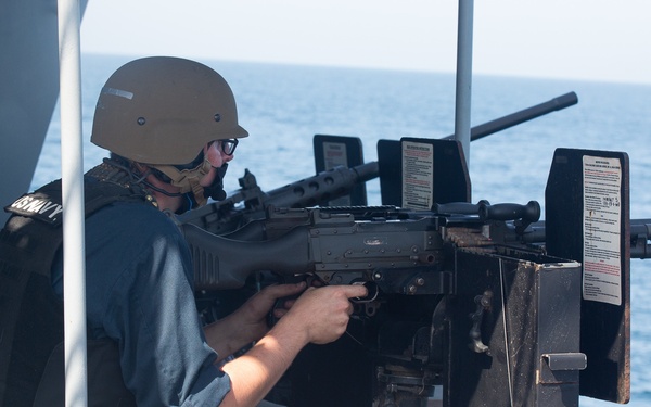 A naval personnel member wearing a tan helmet, glasses, and body armor is operating a mounted machine gun on the deck of a naval vessel at sea. A spent brass casing is visible ejecting from the weapon, indicating active firing. A second crew member in dark uniform is visible in the background.