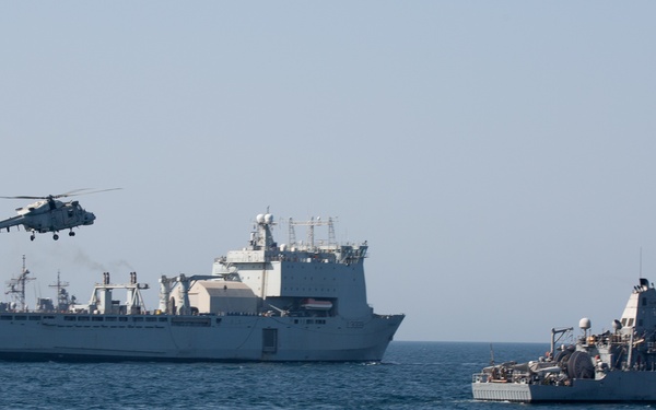 A naval scene showing multiple military vessels at sea with a helicopter in flight. The image depicts what appears to be a multinational naval operation or exercise, with at least two large warships and a helicopter operating in close proximity. An American flag is visibly flying from one of the vessels on the right side.