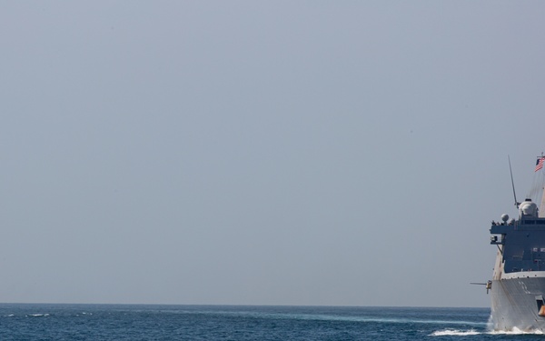 Two naval vessels are underway on open blue water under a hazy sky. The foreground vessel is a large US Navy amphibious transport dock ship, identifiable by its distinctive boxy superstructure and an American flag flying from its mast. A smaller warship is visible in the background to the left, appearing to fly a different national flag.