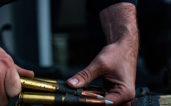 A person is handling large-caliber ammunition rounds arranged in a belt or linked configuration on a dark surface. The rounds appear to be a mix of brass-cased cartridges with copper and white-tipped projectiles, consistent with machine gun or heavy weapon ammunition. The hands are actively manipulating or organizing the linked ammunition chain.