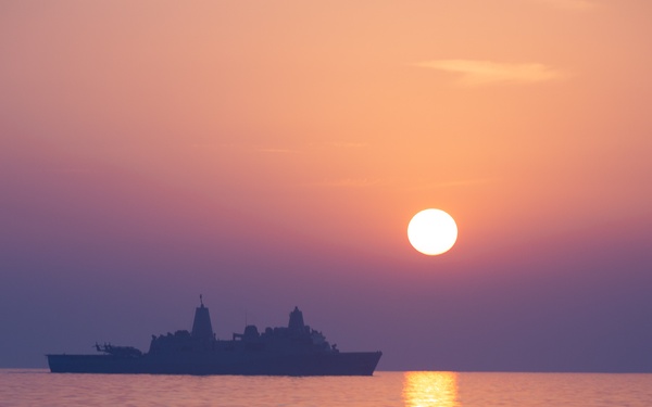 A large naval warship is silhouetted against a vivid sunset over calm open water. The sun is positioned just above the horizon, casting a warm orange and golden reflection across the sea's surface. The sky transitions from deep purple-blue tones in the upper frame to warm orange and pink near the horizon.