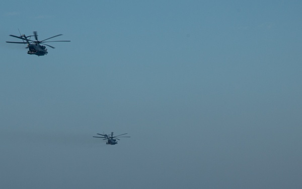 Two military helicopters fly in formation over open ocean near a large naval vessel. The ship appears to be a US Navy amphibious transport dock (LPD-class), identifiable by its distinctive superstructure and flat deck sections. The scene suggests a military exercise or operational deployment at sea.