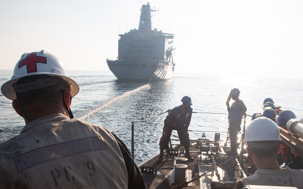 Naval personnel aboard a patrol craft (marked 'PC 9') are conducting an underway replenishment (UNREP) operation with a larger supply vessel visible in the background. A crew member in the foreground wears a white hard hat and vest marked with red crosses, indicating a medical/safety role. A transfer line or hose is visibly stretched between the two vessels during the at-sea replenishment evolution.