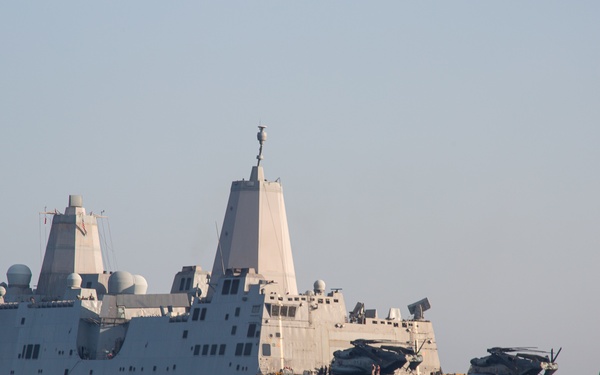 A large US Navy amphibious transport dock ship is underway at sea, with a floating dock or causeway section visible alongside its stern carrying military helicopters. The vessel features the distinctive angular stealth superstructure characteristic of San Antonio-class (LPD-17) amphibious ships. A small rigid inflatable boat (RIB) is visible in the water nearby, likely conducting escort or security duties.