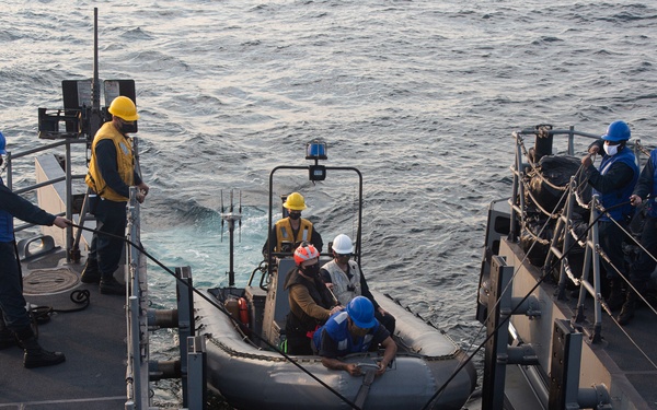 Naval personnel are conducting small boat operations on the deck of a military vessel at sea. A rigid inflatable boat (RIB) is secured on deck while crew members in safety gear work around and inside it. The scene appears to depict a boat recovery or launch operation with multiple personnel in assigned roles.