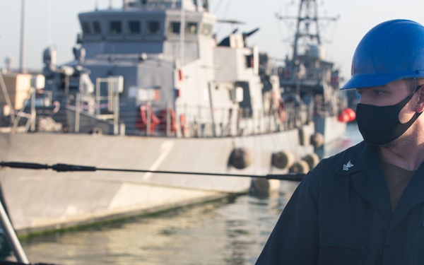 A uniformed naval sailor wearing a blue hard hat and black face mask is stationed on the deck of a vessel at a harbor, handling mooring lines or rigging. The sailor's uniform displays name tapes and rank insignia consistent with the U.S. Navy. Several gray naval vessels are docked in the background at what appears to be a military port facility.